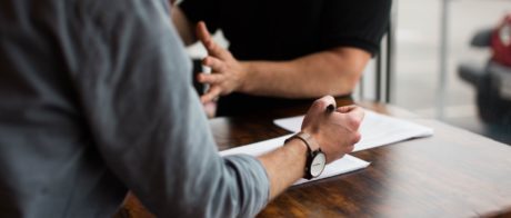 Side view of a person in a meeting who's holding a pen above a piece of paper on a desk.