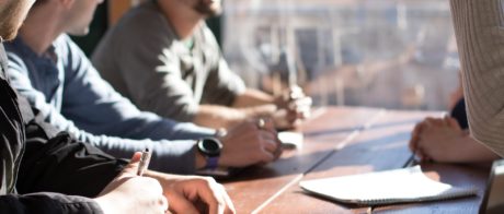 People in a meeting, sitting at a desk with pens and paper.