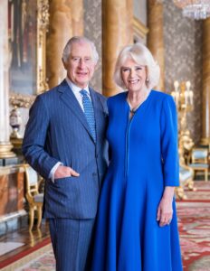 King Charles III in a blue pin-striped suit with a blue tie standing and smiling with Queen Camilla who is smiling and wearing a blue dress. 