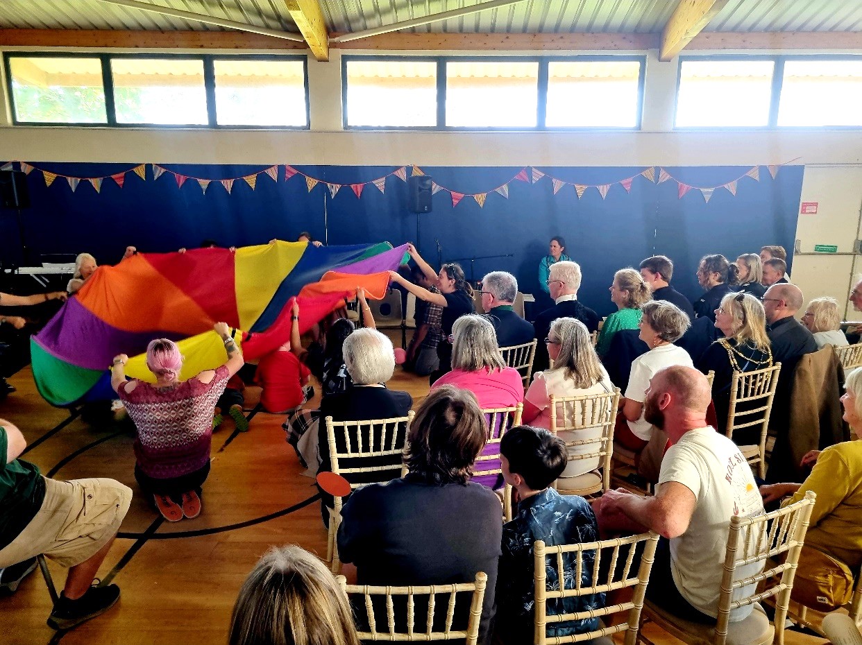 A young group of people playing with a multi-coloured parachute with an audience watching